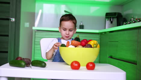 little caucasian boy in jeans romper sits at table in the kitchen. toddler pulls a bowl of veggies closer and picks an eggplant.