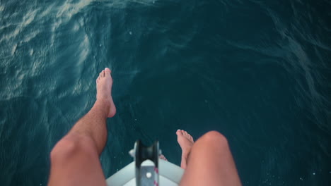 Man-sitting-on-the-bow-of-a-boat-dangling-his-feet-over-the-clear-blue-ocean-water