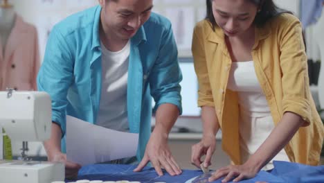 diseñadores masculinos y femeninos asiáticos con máquina de coser ayudándose mutuamente crea una plantilla del vestido futuro. diseñadores ayudándose a cada uno a dibujar un contorno de tiza de los detalles