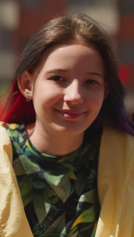 positive girl with colored strands of hair sits on swing in yard near high-rise building. happy child smiles sweetly looking at camera closeup on blurred background