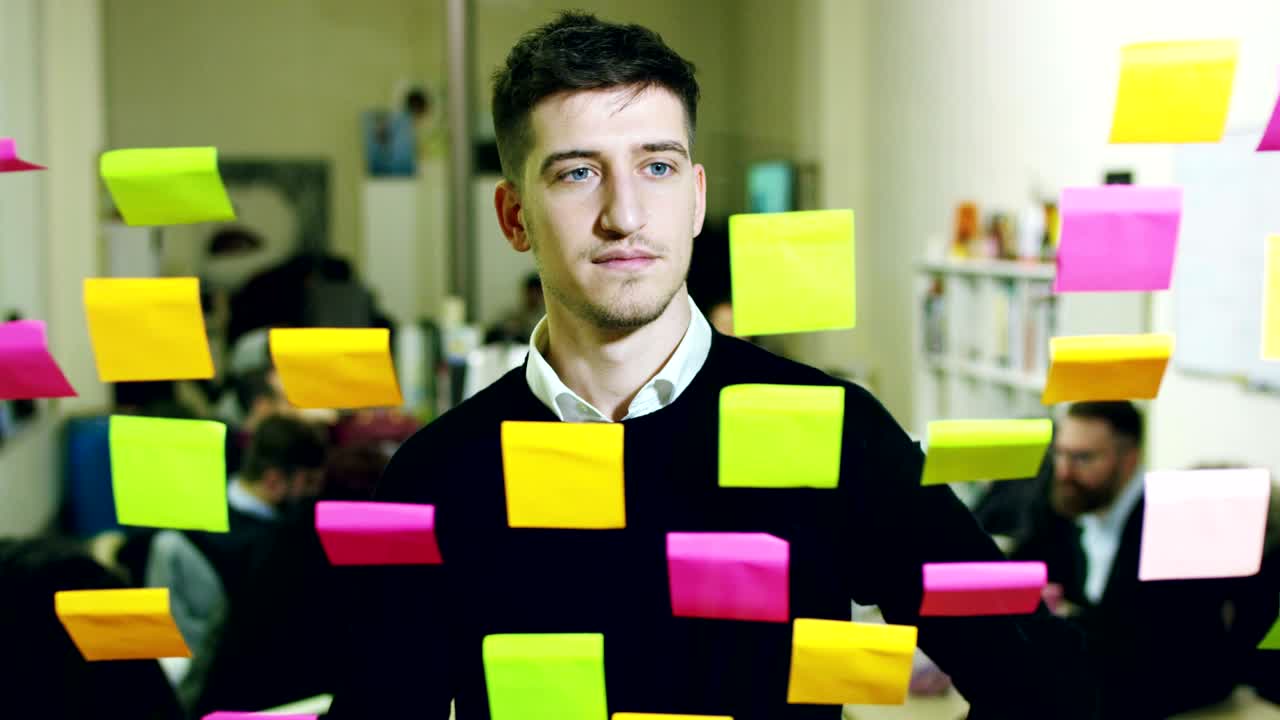 retrato de un hombre sonriente en una agencia reflexiva y creativa con notas coloridas y planificación colgando detrás del vidrio y la gente trabajadora.