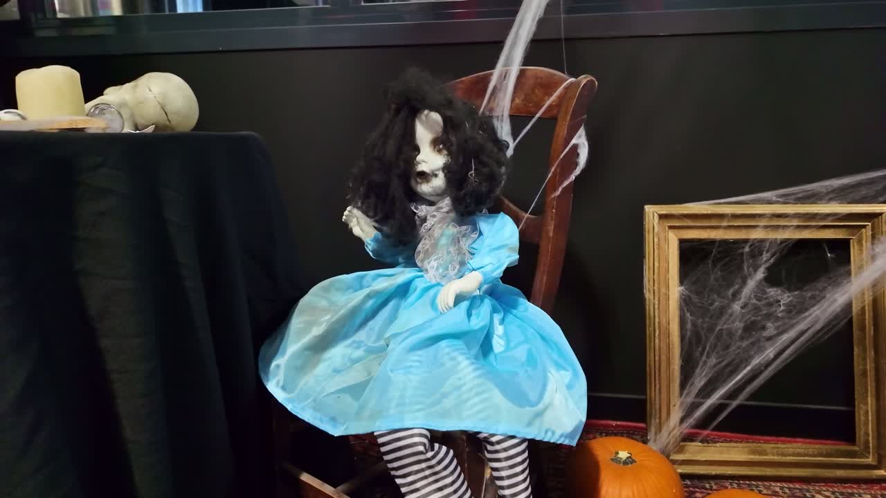 Creepy haunted doll with messy hair and blue dress sits on chair, spooky Halloween decoration with spiderwebs, pumpkins, and skull. Close-up