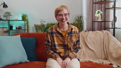 pretty woman smiling friendly at camera, waving hands gesturing hello, hi, greeting at home on sofa