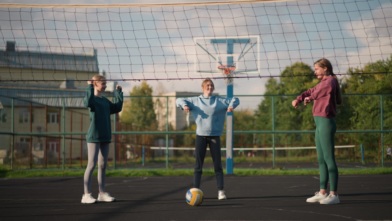 спортивные женщины стоят на открытом волейбольном корте, тренируются, поворачивая руки, волейбол лежит впереди, с спортивным сооружением и жилыми зданиями на заднем плане