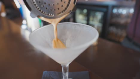 Espresso-martini-poured-into-chilled-glass-from-shaker-on-bar-counter,-close-up