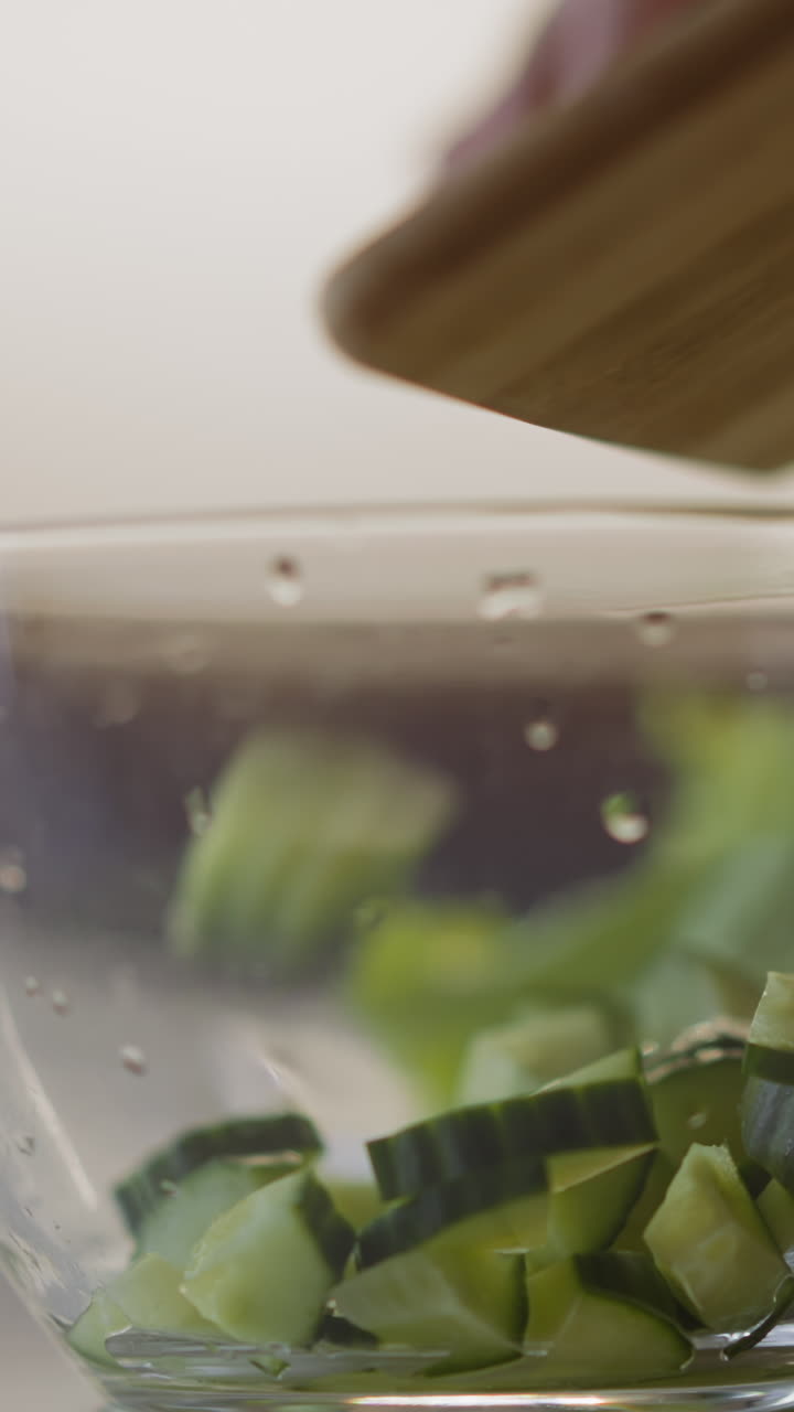 Pouring juicy cucumber slices into glass bowl from board cooking healthy salad on table in light kitchen extreme close view slow motion