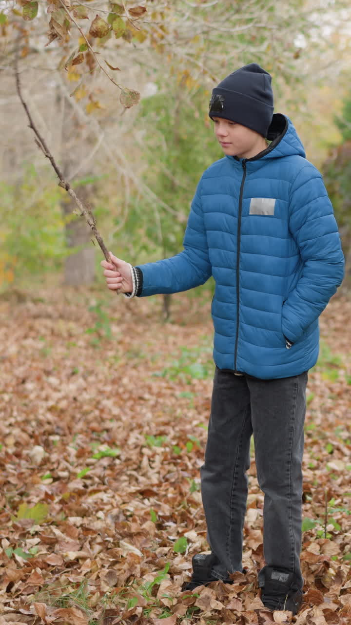 파란색 재과 검은색 모자를 입은 아이의 클로즈업, 나무 바닥에 건조한 가을 잎을 막대기로 놀이롭게 어, 활기차고 즐거운 가을 야외 장면을 만니다.
