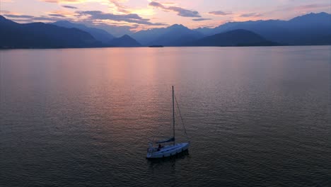 Aerial-View-Of-A-lone-sailboat-glides-peacefully-on-a-tranquil-lake-surrounded-by-mountains,-with-a-spectacular-and-vibrant-sunset-sky-reflecting-on-the-water