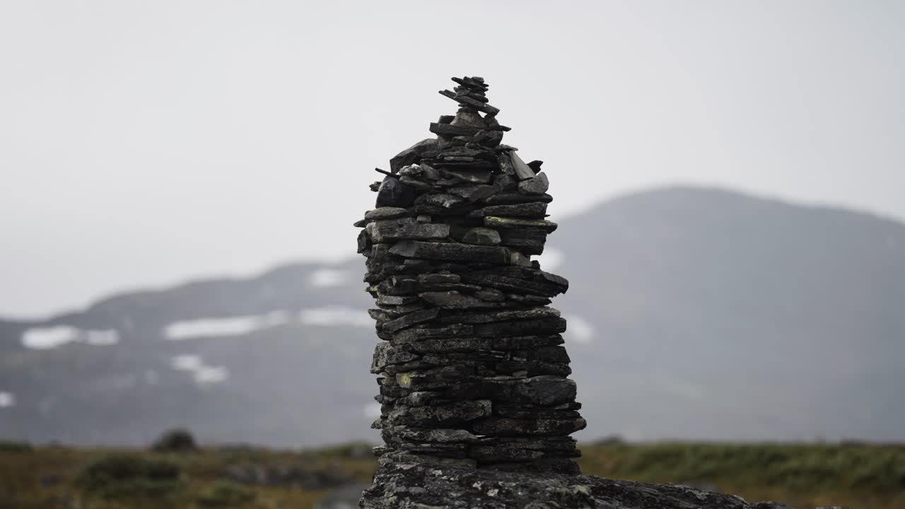 A dark stone cairn in the bleak northern landscape. Misty snow-covered mountains in the background. Parallax shot.