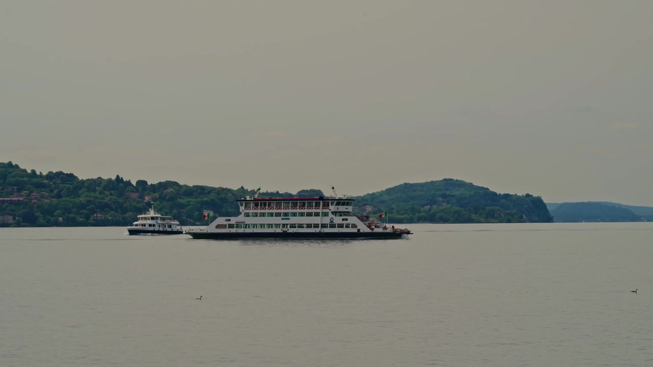 Rekaman pelacakan sinematik sebuah feri yang berlayar melintasi danau Italia dengan air tenang, garis pantai yang jauh, dan langit mendung abu-abu yang murung menciptakan suasana perjalanan yang tenang.