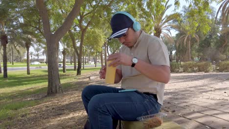 a man drinking water, sitting on a bench with a backpack and a cell phone