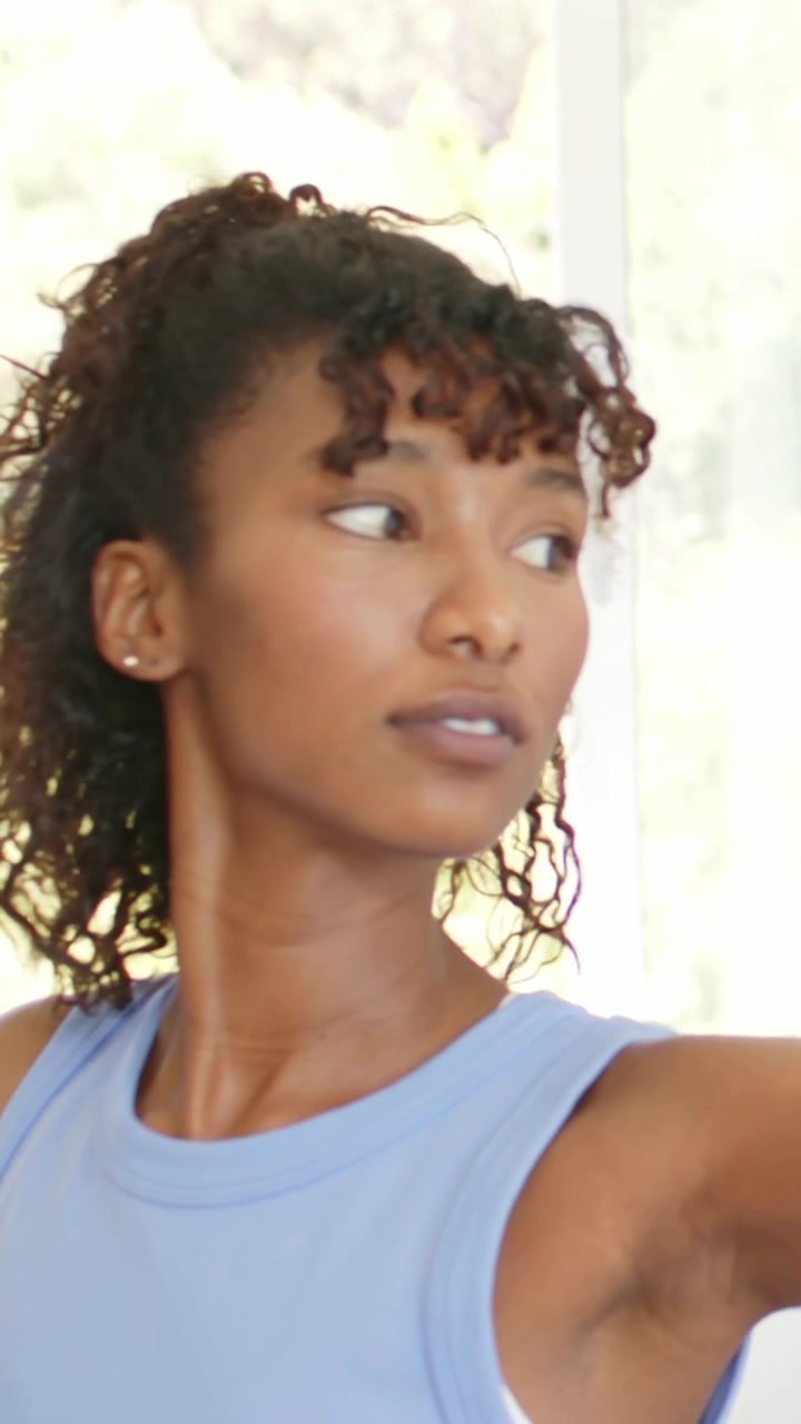 Vertical video: Starting woman in blue top holding arm, rotating and turning head by window, watch