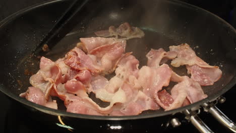 bacon cooking in non-stick skillet on stovetop is stirred with tongs