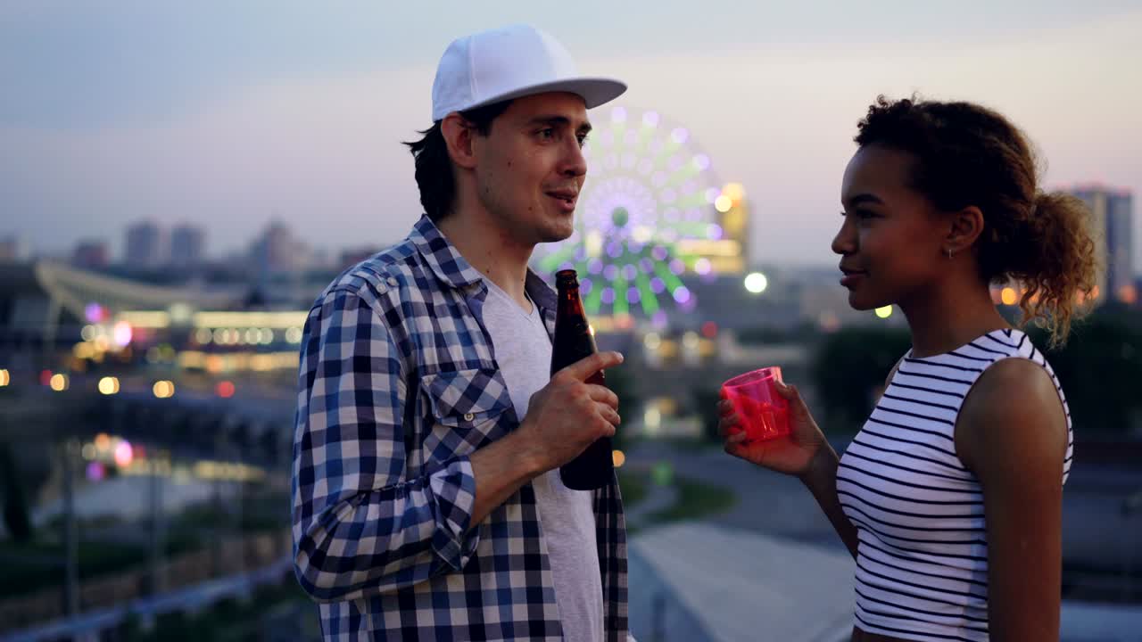 Pretty African American girl is talking to her Caucasian friend and drinking at rooftop party in the evening in summer enjoying conversation and beautiful view.