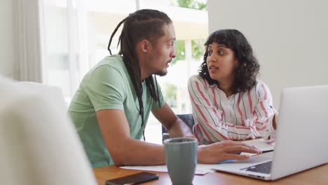 hombre biracial con dreadlocks y pareja femenina usando computadora portátil juntos y hablando en la sala de estar