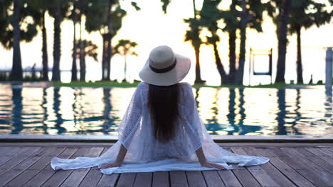 Rückseite-Einer-Stilvollen-Frau-Mit-Sommerhut,-Die-Am-Pool-Sitzt-Und-Das-Sonnenlicht-Der-Goldenen-Stunde-Am-Meereshorizont-Beobachtet