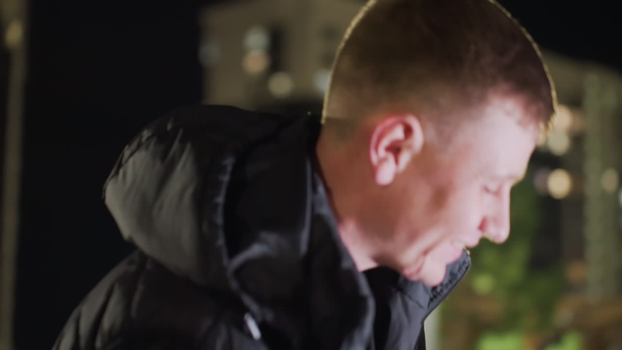 White man walks toward woman seated outdoors at night and kisses her lovingly as streetlight shines warmly on them in romantic city setting surrounded by tall buildings and soft glowing lights