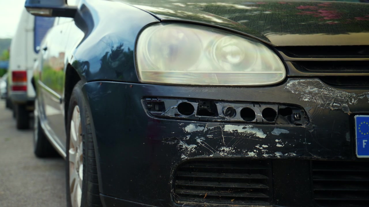 Auto nera che mostra danni frontali significativi, con paraurti rotto, vernice graffiata e faro appannato