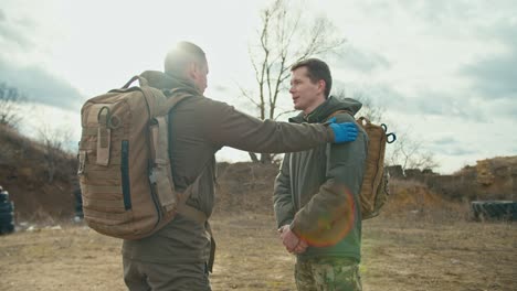 Side-view-of-a-confident-military-man-in-a-dark-green-uniform-and-with-a-backpack-on-his-shoulders-pats-his-colleague-and-partner-on-the-shoulder-as-a-sign-of-support-outside-the-city