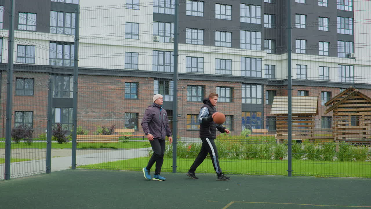 two white men walking with basketball on fenced urban court near apartment buildings, playful pickup session, casual jackets and sneakers, light dribble and friendly banter, green lawn and playground