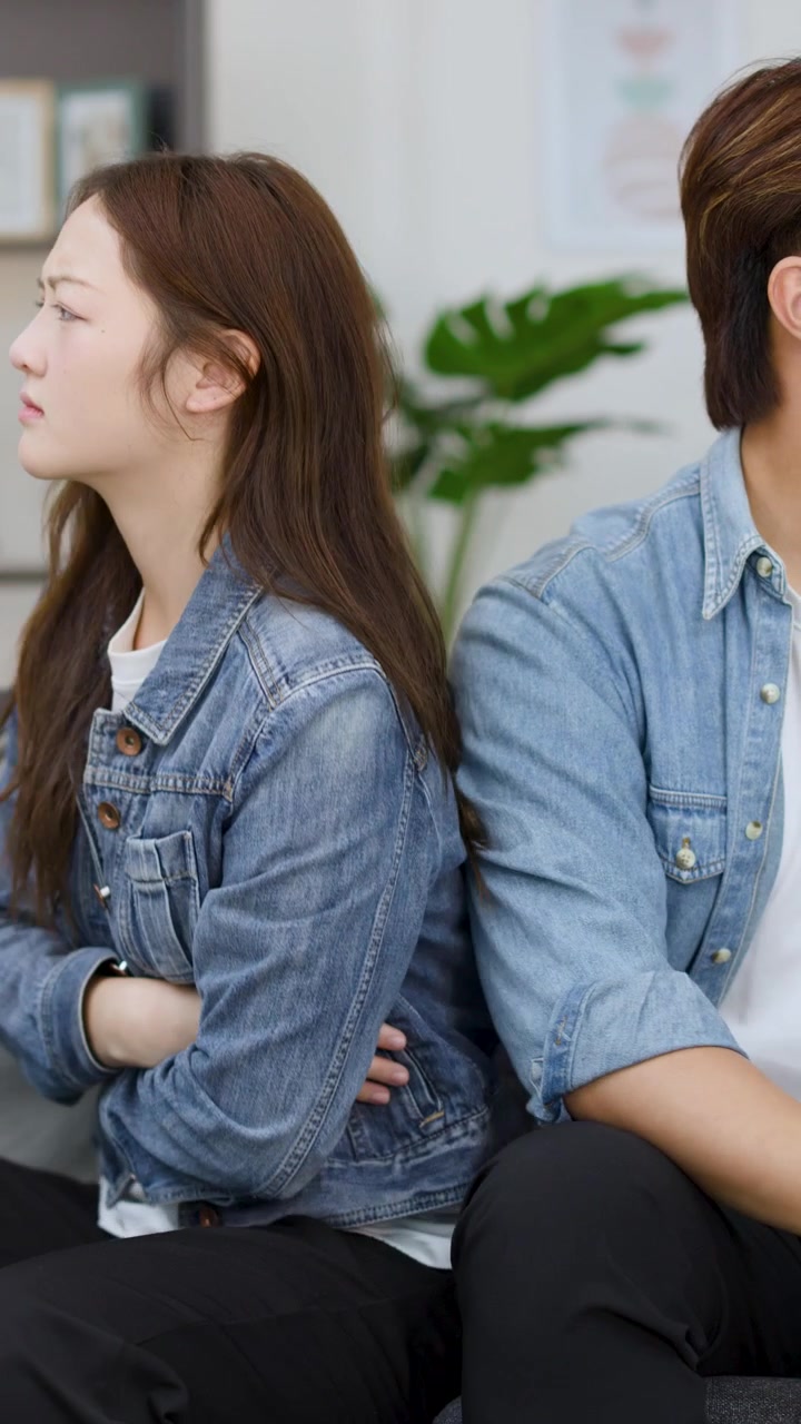 An upset young couple sits on a sofa, arguing and avoiding eye contact indoors