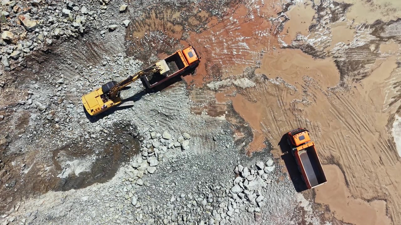 Granite quarry site with trucks and excavator working on rocky ground near a dusty road during daytime
