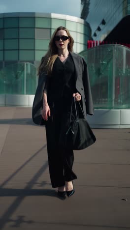 vertical - stylish caucasian businesswoman in a black suit and sunglasses walks confidently on an outdoor terrace of a modern office building in the city - slow motion, tracking pull back