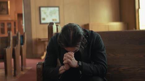 Sad,-depressed,-emotional,-young-man-in-black-suit-praying-in-church-pew-in-cinematic-slow-motion-with-folded-hands