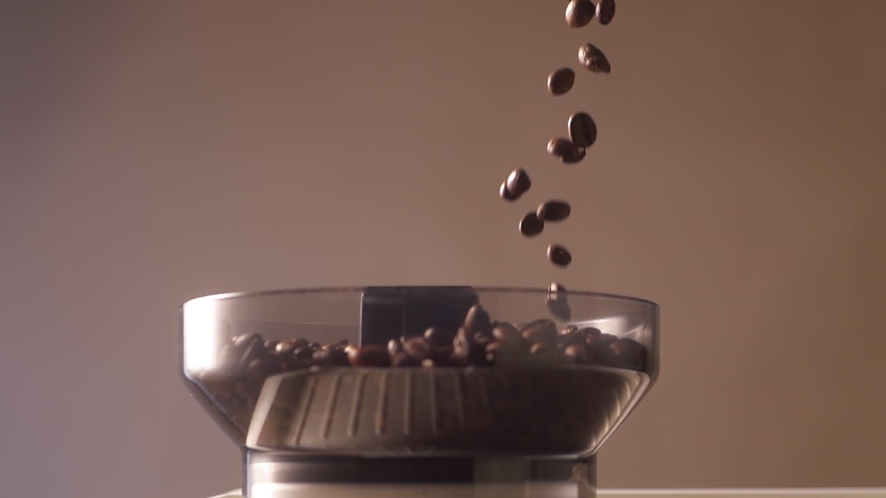 Un flusso di chicchi di caffè tostati viene versato nella tramoggia trasparente di un macinacaffè per espresso. La ripresa al rallentatore enfatizza la consistenza dei chicchi, i toni ricchi e la precisione della preparazione del caffè.