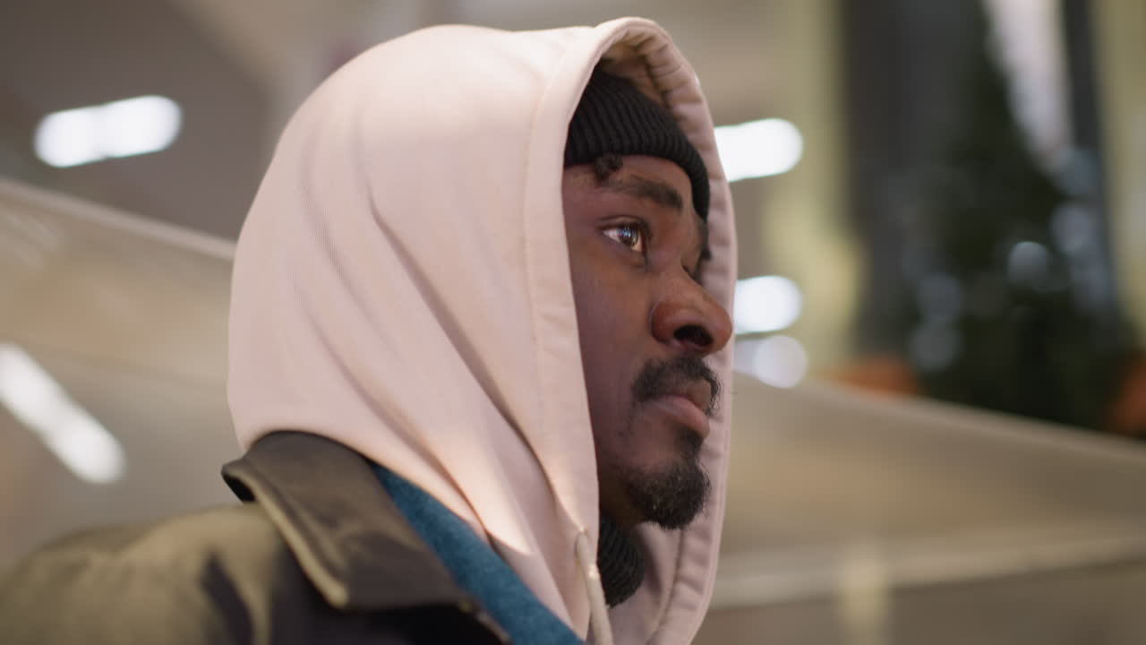 A young man wearing a hoodie and beanie stands thoughtfully in a modern shopping mall. His calm expression and subtle gaze are framed against soft indoor lighting in the commercial space