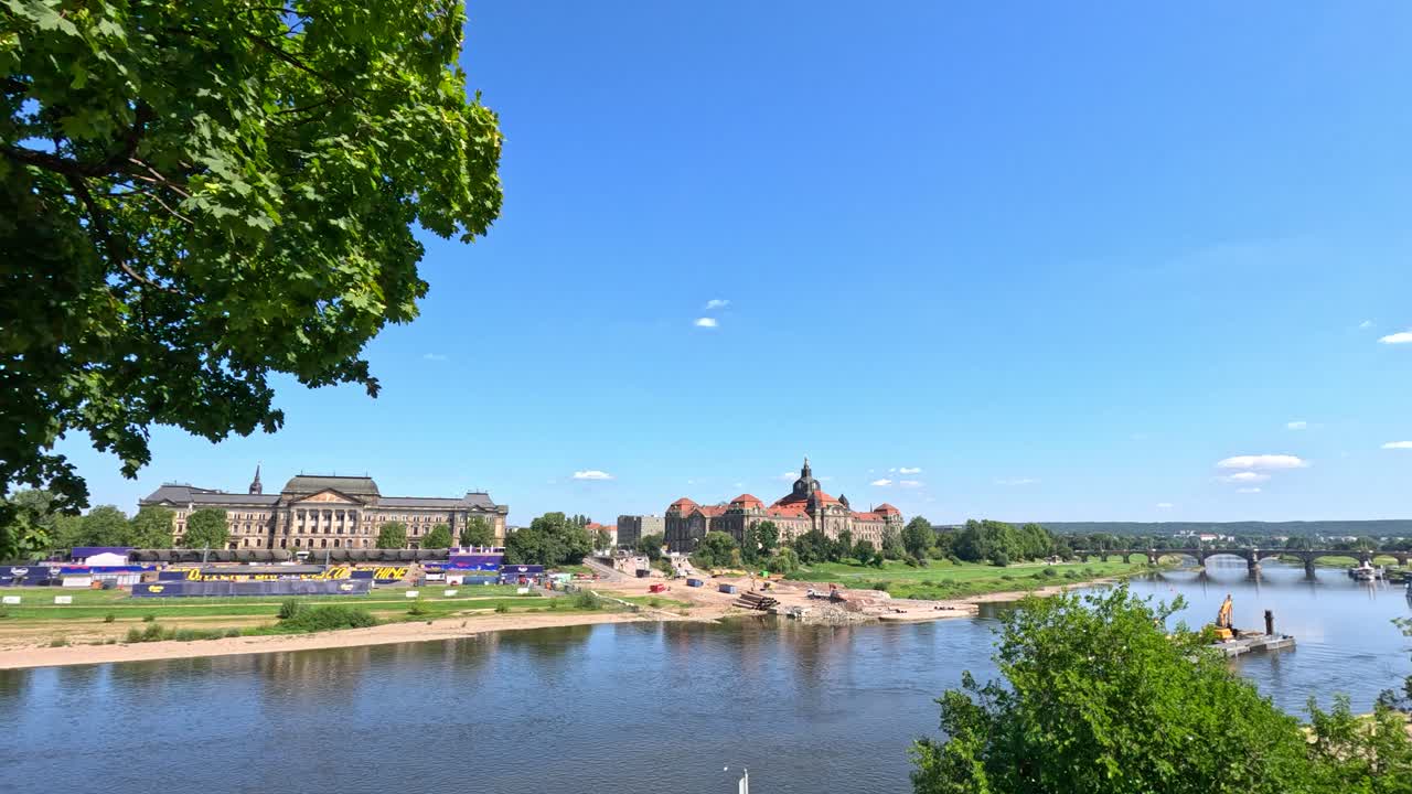 Camera pans along sunny Dunkirk riverside, showcasing historic buildings, leafy trees, and calm water