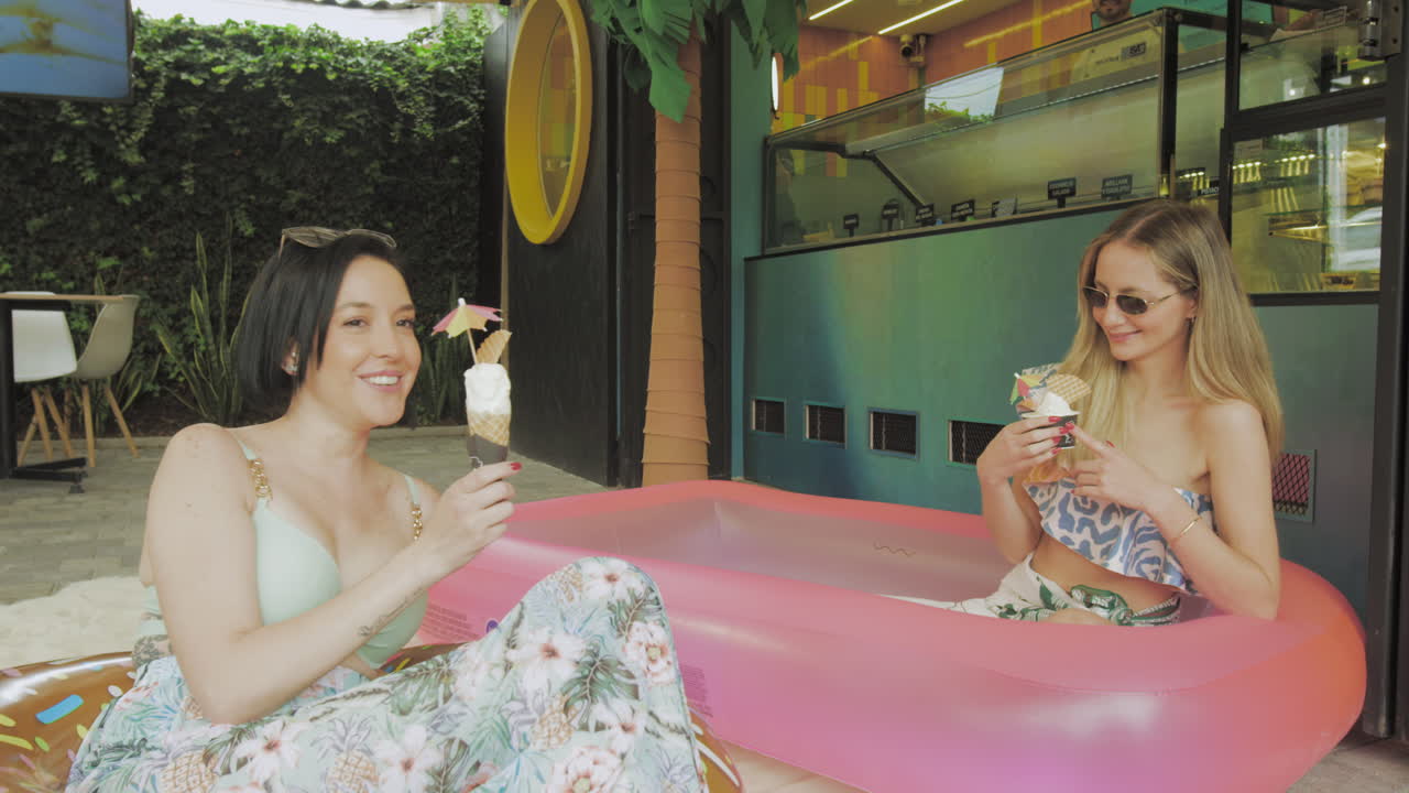 Two young women enjoy ice cream while relaxing in an inflatable pool in front of a modern ice cream parlor with tropical decor.
