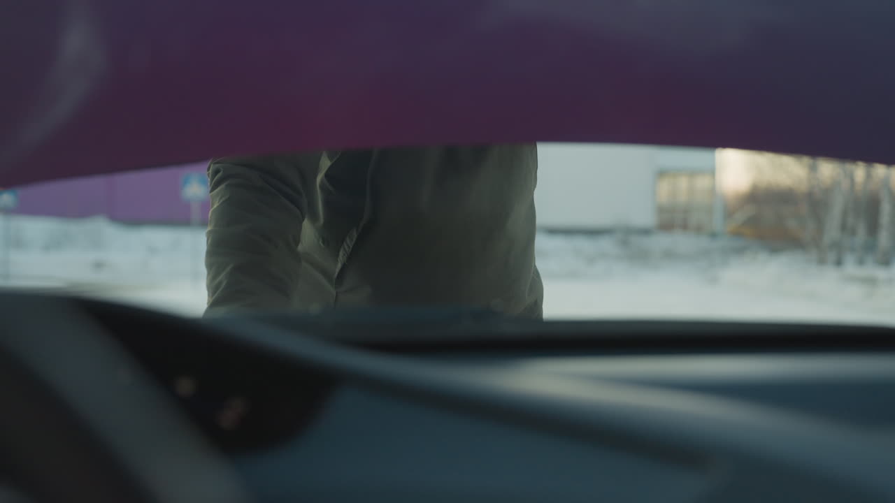 Close up from inside vehicle shows person removing support rod and closing car bonnet after engine check in cold winter environment with snow-covered ground and distant buildings in background