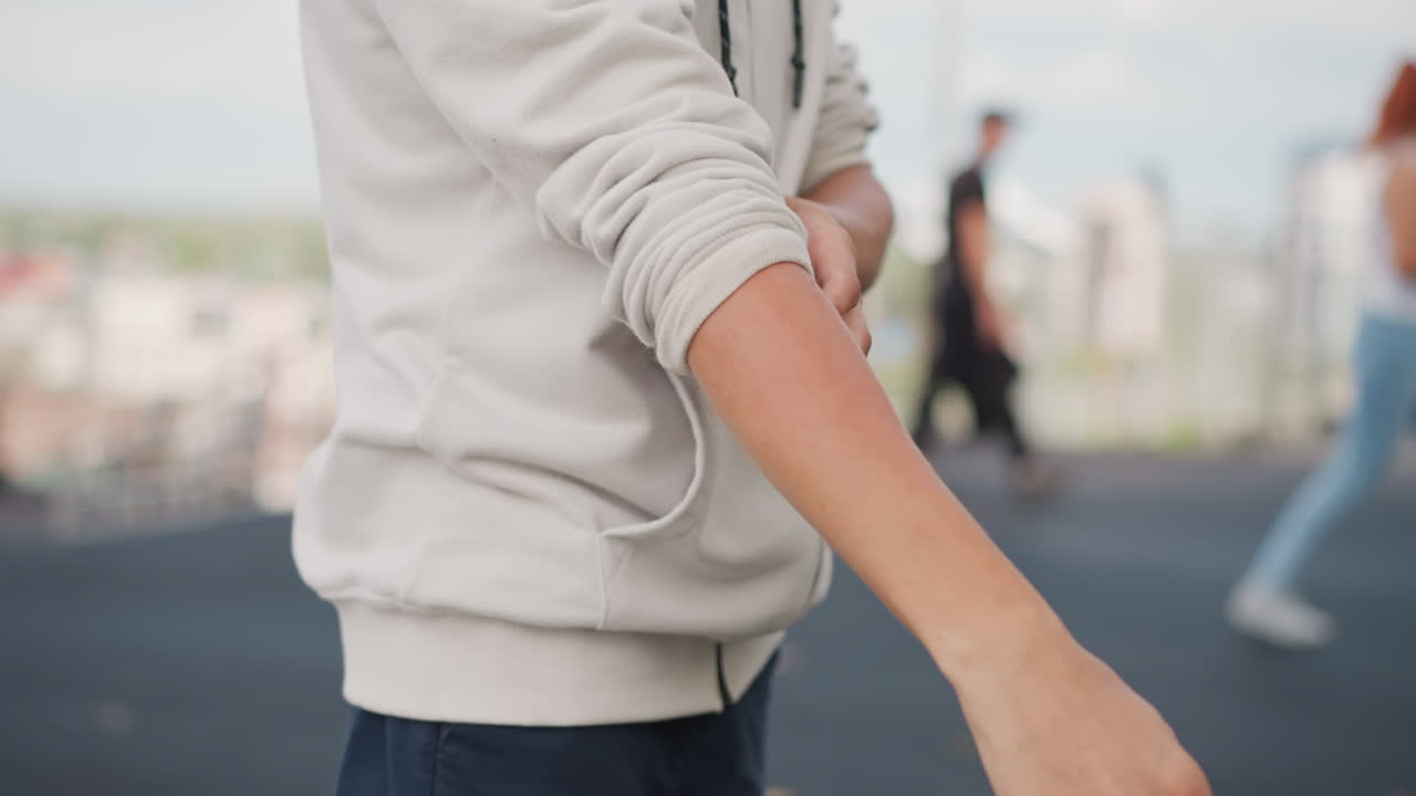 도시 산책로에서 소매를 정리하는 남성, 후드티와 편안한 자세, 멀리 보이는 보행자와 스카이라인, 부드러운 흐린 조명, 일상적인 도시 풍경, 천 질감과 자연스러운 클로즈업