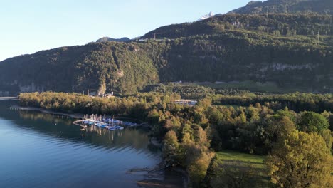 Una-Hermosa-Vista-Panorámica-Sobre-El-Lago-Walensee-Con-Aguas-Serenas-Y-Azules,-Un-Pequeño-Puerto-Deportivo-Con-Veleros-Y-árboles-Bañados-Por-El-Sol-En-Las-Colinas-De-Las-Montañas