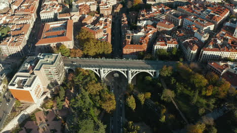 Fly-above-historic-road-arch-bridge-Segovia-Viaduct-spanning-valley-in-city.-Cars-driving-in-streets.