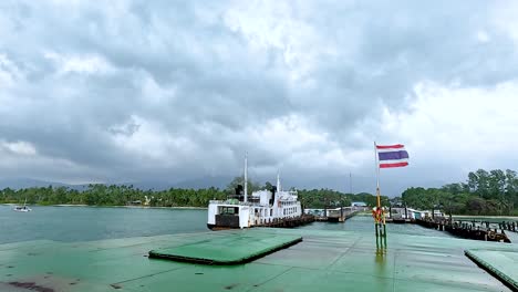 a ferry approaches the dock in koh samui, thailand, under overcast skies. the thai flag waves prominently in the foreground