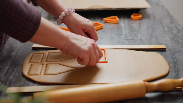 Un panettiere utilizza un taglierino arancione per creare forme precise su una pasta marrone stesa; nelle vicinanze, altri taglierini per dolci e un matterello di legno sono appoggiati su un piano di lavoro ruvido