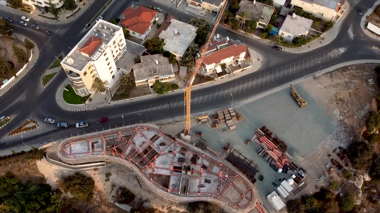 Aerial view shows ongoing construction of a large building in a bustling city. Surrounding roads and existing structures highlight the urban environment and development efforts