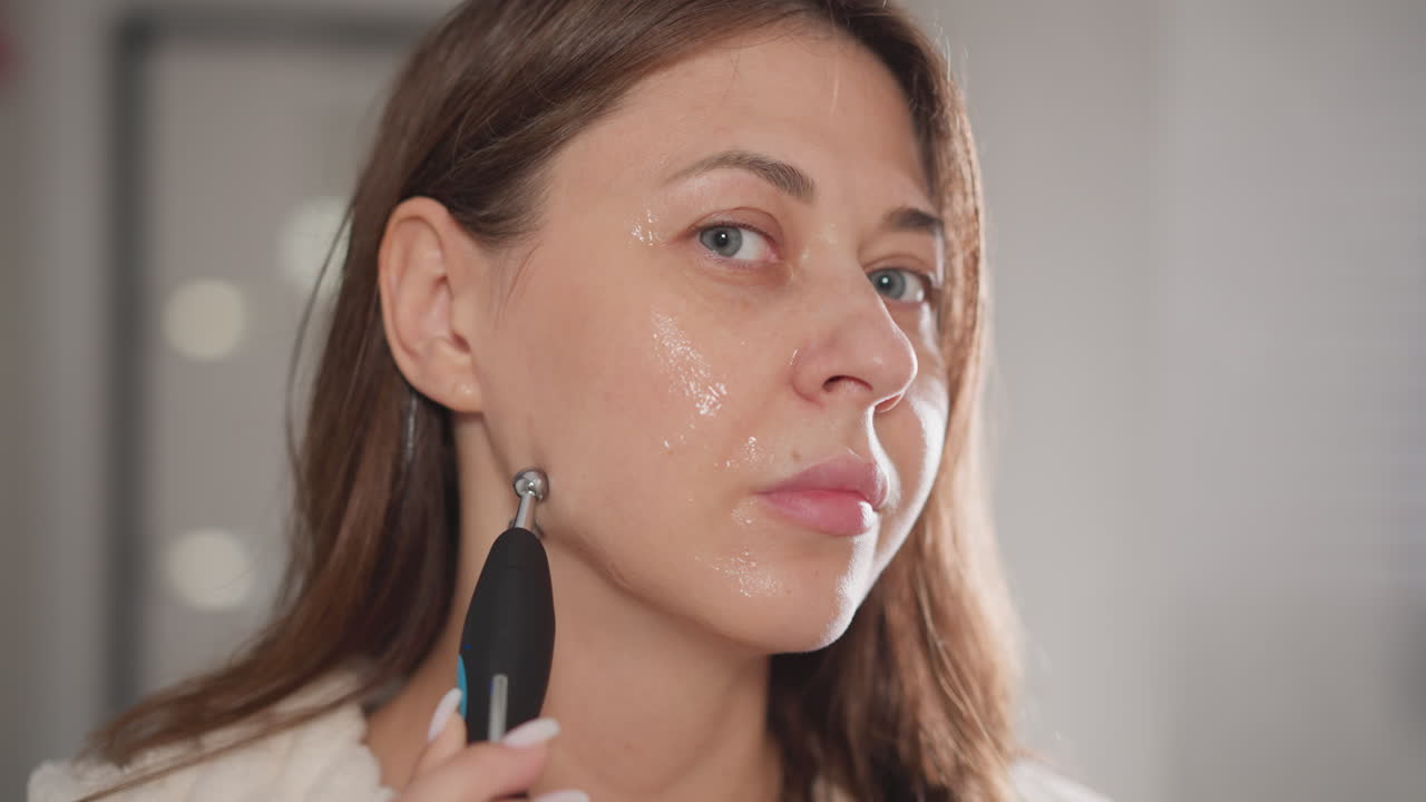 mujer masajea áreas problemáticas de la cara con una herramienta de microcorriente. mujer morena señala un dispositivo de masaje sobre la cara en el baño en el fondo en cámara lenta