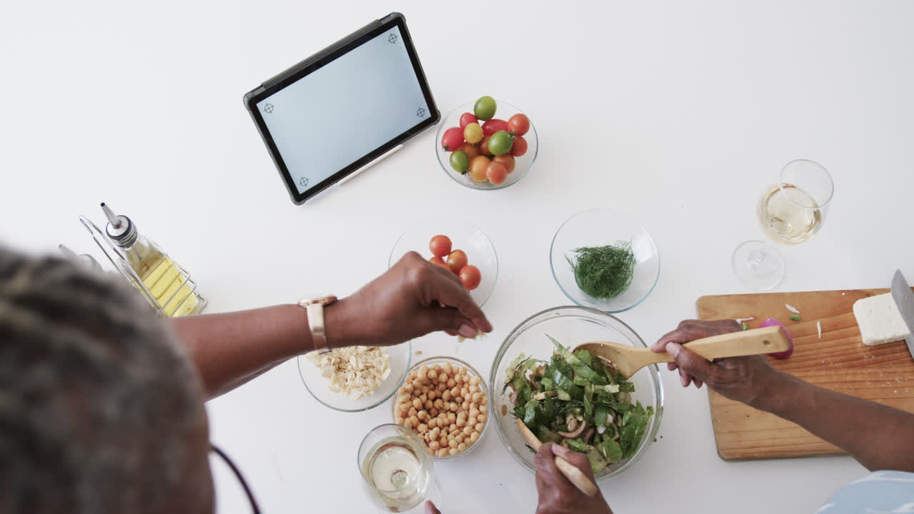 Senior african american female friends making salad, using tablet with copy space, slow motion