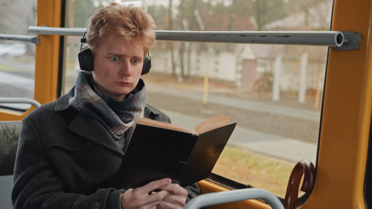 Homme blanc lisant dans un bus, écrivain créateur étudiant des pages avec des écouteurs et une écharpe, expression concentrée, vue rapprochée par la fenêtre d'un itinéraire de banlieue, ambiance littéraire calme pendant le trajet matinal.