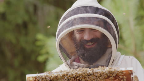 Smiling-happy-beekeeper-in-bee-suit-inspects-hive-frame-covered-in-honeybees