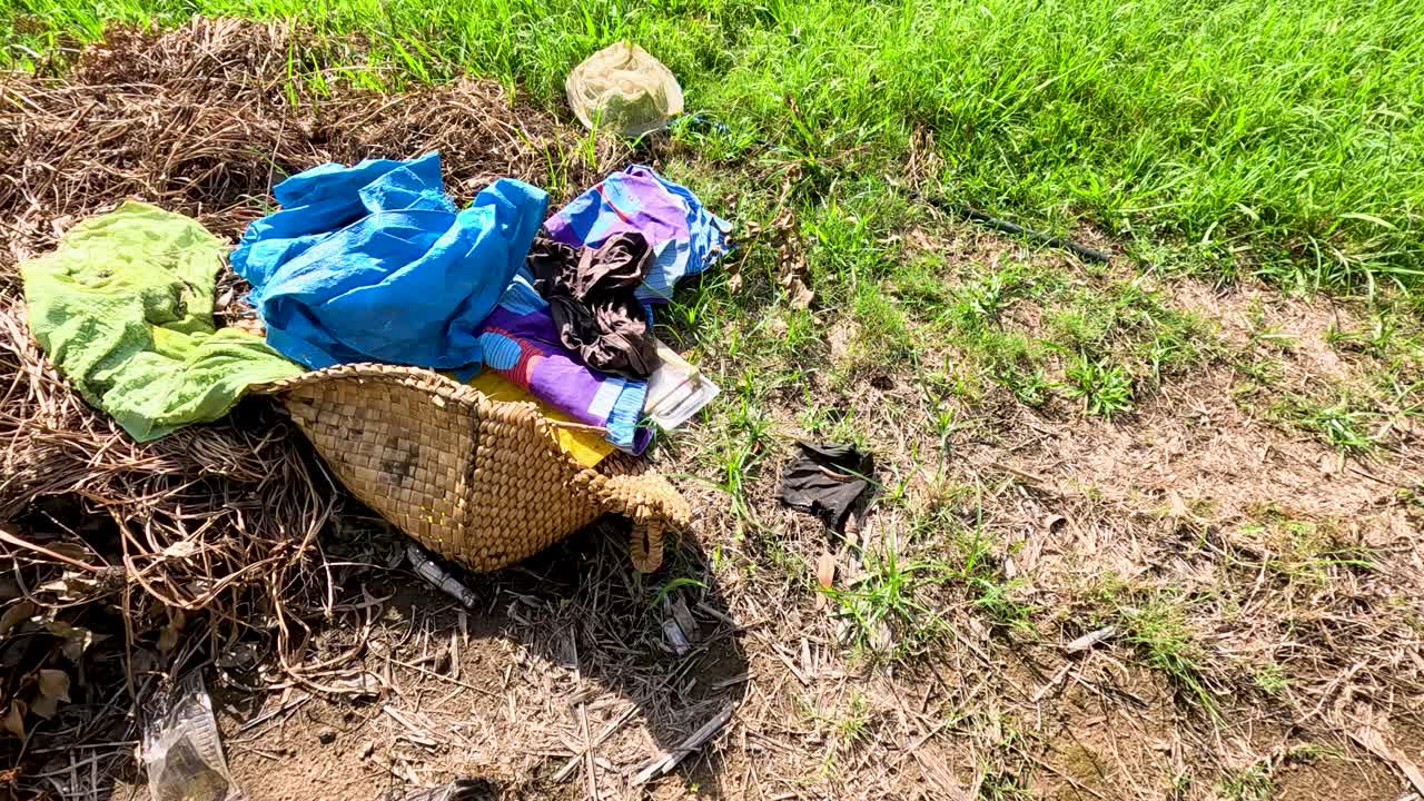 Laundry basket with clothes and rubbish in sunlit rural field, camera pans across polluted area