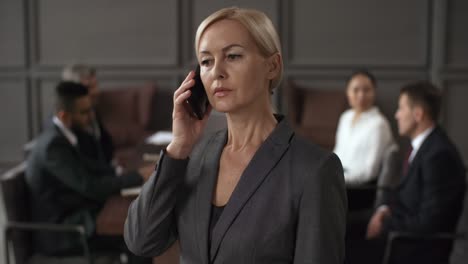 portrait of a serious businesswoman standing and speaking on mobile phone, while in blurred background group of business executives sitting around table during meeting