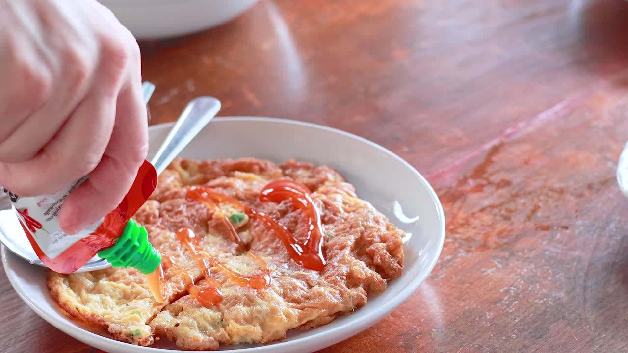 한 손은 태국 스타일의 오멜 위에 칠리 소스를 부어 맛을 높인다. 자연 조명이 있는 나무 테이블 위에 장면이 설정되어 있다.