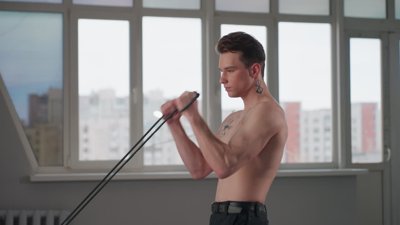 shirtless man stands, grips heavy rope handles, braces body and pulls rope downward in intense strength training move with residential buildings visible through large window behind him
