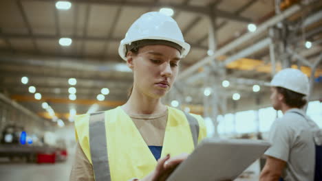 female factory worker in safety vest and hard hat using digital tablet