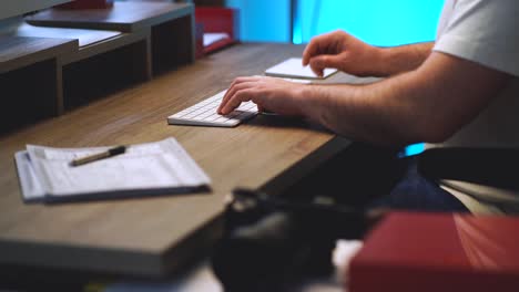 creative person sitting at his home office modern wood desk typing on a white wireless computer keyboard, clicking a mouse trackpad and writing down notes on notepad with a blue light in background