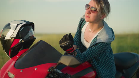 a young woman adjusts her sunglasses while looking at her reflection in the motorcycle mirror, she seems thoughtful, with the soft backdrop of an open field and the bike s red body